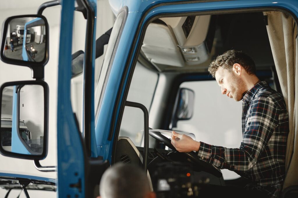 A male mechanic in a plaid shirt reads a checklist inside a truck cab, focused and serious.